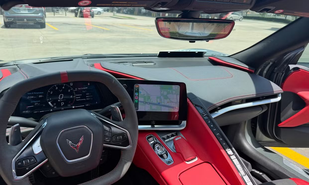 Corvette Stingray cockpit view showing the steering wheel, digital gauge cluster, and vertical infotainment display with red leather accents